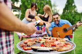 man's hand holds a barbecue tongs with a juicy delicious meat steak against the background of a barbecue grill with meat and vegetables and a group of friends on a picnic who are having fun and