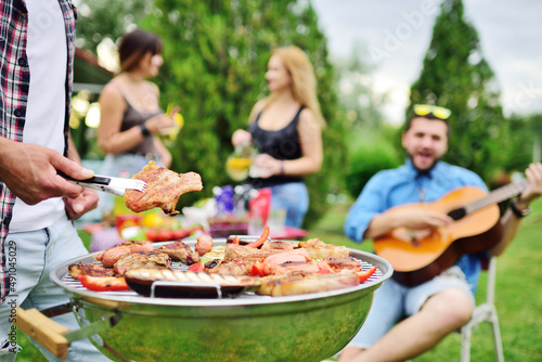 man s hand holds a barbecue tongs with a juicy delicious meat steak against the background of a barbecue grill with meat and vegetables and a group of friends on a picnic who are having fun and