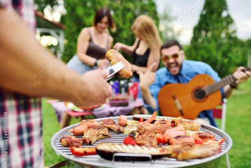 man's hand holds a barbecue tongs with a juicy delicious meat steak against the background of a barbecue grill with meat and vegetables and a group of friends on a picnic who are having fun and