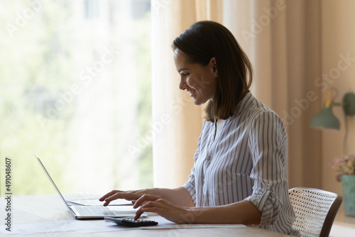 Smiling young woman sit at table do accounting work from home office, making calculations on calculator, pay utility bills use laptop. Distancing job, manage family budget, finances control concept