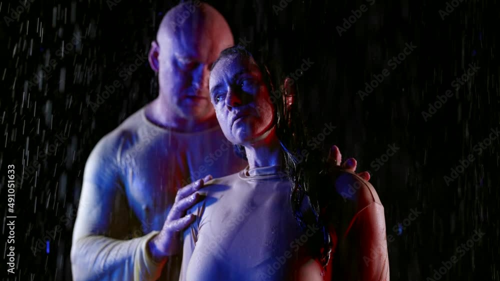 An attractive bald man embraces his beloved in the rain. A happy couple enjoys each other. A love story in raindrops. Shot in a dark studio. Slow motion. An attractive bald man embraces his beloved in the rain. A happy couple enjoys each other. A love story in raindrops. Shot in a dark studio. Slow motion.