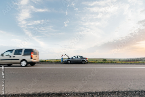 A young girl stands near a broken-down car in the middle of the highway during sunset and tries to call for help on the phone. Waiting for help. Car service. Car breakdown on road. © Andrii