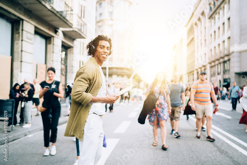 Half length portrait of cheerful male blogger in bluetooth headphones crossing city road and smiling at camera during vacations journey, happy hipster guy with modern touch pad listening audio guide