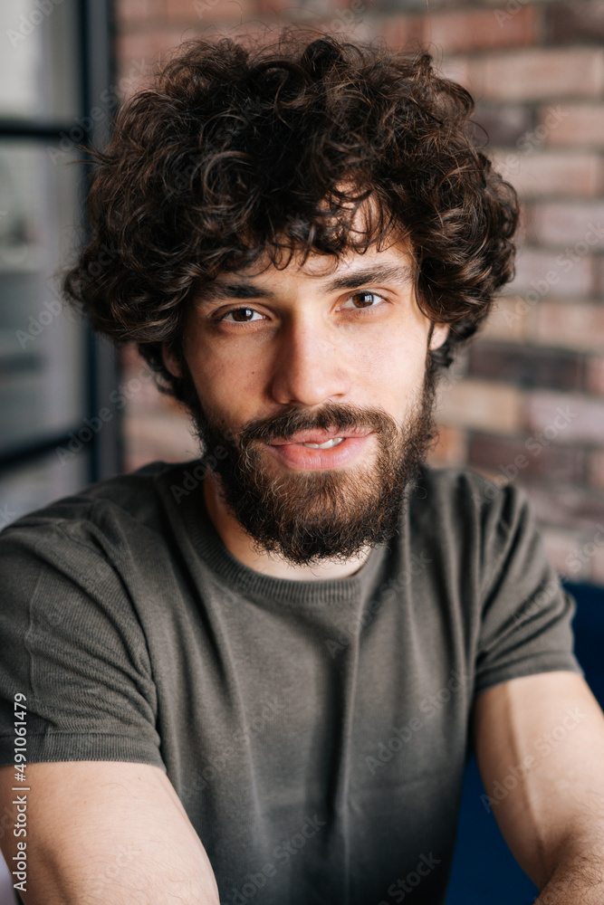 Close-up vertical portrait of friendly handsome young man looking at camera sitting on chair in cozy living room with brick wall. Front view of confident curly male posing at home. Close-up vertical portrait of friendly handsome young man looking at camera sitting on chair in cozy living room with brick wall. Front view of confident curly male posing at home.