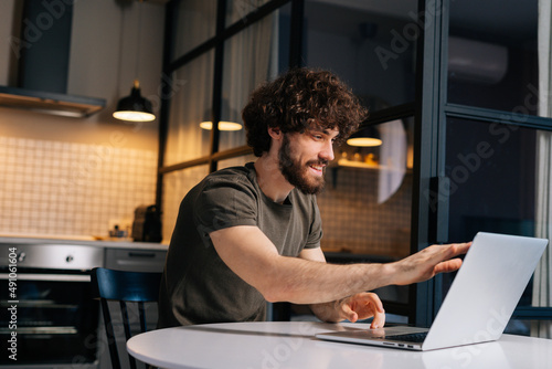 Side view of smiling young freelancer male opening laptop and starts typing on keyboard sitting at table in kitchen with modern interior. Smiling businessman working remote at home office.