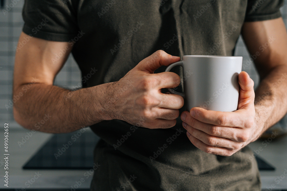 Close-up cropped shot of unrecognizable man holding in hand cup with morning coffee, standing in kitchen with modern light interior, enjoying fresh warm drink at home office. Close-up cropped shot of unrecognizable man holding in hand cup with morning coffee, standing in kitchen with modern light interior, enjoying fresh warm drink at home office.