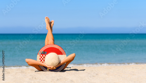Fototapeta Naklejka Na Ścianę i Meble -  A slender girl on the beach in a straw hat in the colors of the flag of Croatia. The concept of a perfect vacation in a resort in the Croatia. Focus on the hat.