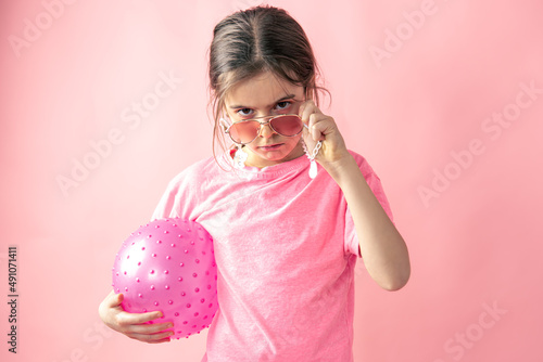 Stylish little girl girl in sunglasses with a ball on a pink background.