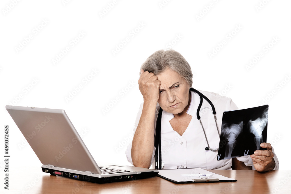 Serious elderly woman doctor sitting at table with computer and looking at x-ray Serious elderly woman doctor sitting at table with computer and looking at x-ray