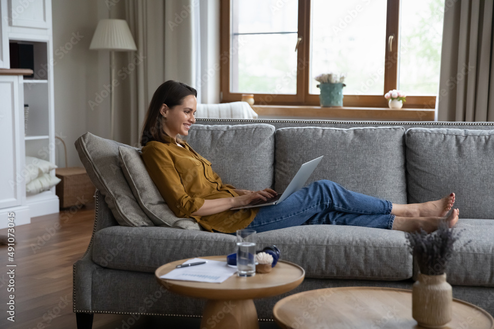 Side view woman sits on sofa in living room put laptop on laps enjoy online chat, share messages in social media, communicates remotely use internet. Modern tech, fun, leisure or telecommuting concept Side view woman sits on sofa in living room put laptop on laps enjoy online chat, share messages in social media, communicates remotely use internet. Modern tech, fun, leisure or telecommuting concept