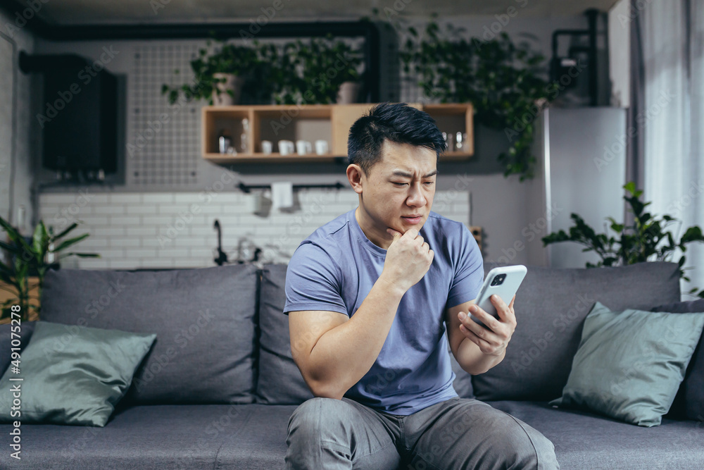Obraz premium Concerned man reading news online looking at phone screen, Asian sitting on sofa at home, serious and sad