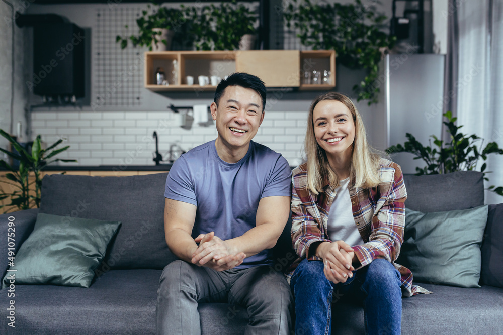 Loving multiracial couple, man and woman, portrait of young family, looking at camera and smiling, sitting on sofa in living room at home Loving multiracial couple, man and woman, portrait of young family, looking at camera and smiling, sitting on sofa in living room at home