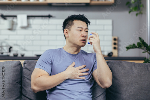 A man with asthma, an Asian man at home sitting on the couch breathing in an inhaler