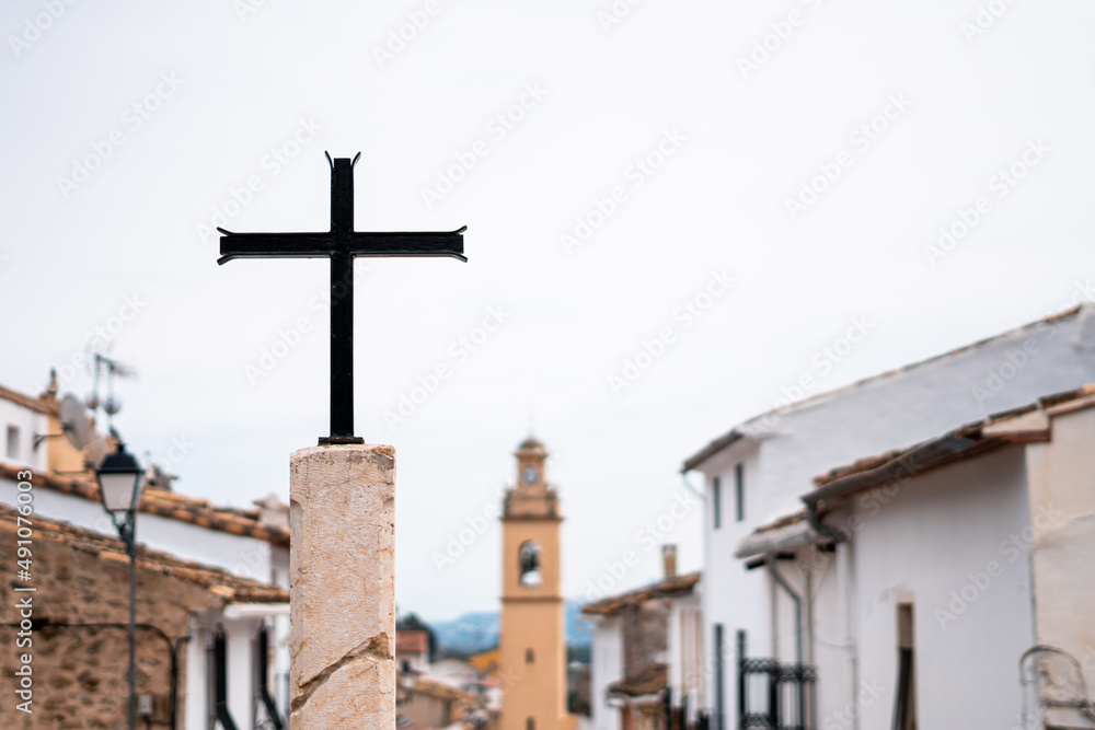 Metallic cross on a stone pedestal, with a village out of focus background.