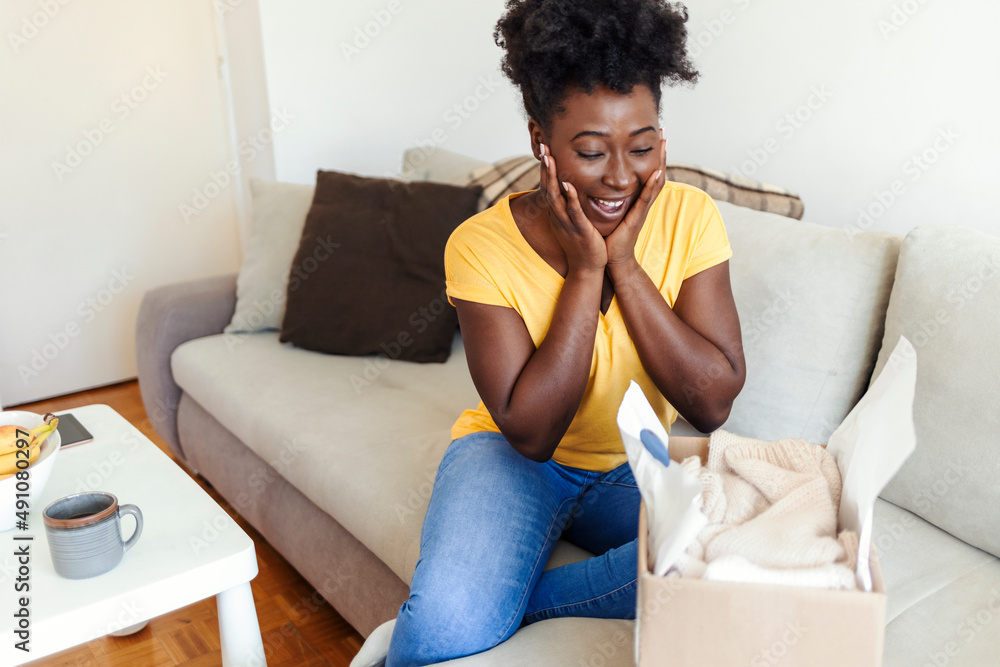 Happy African-American young woman open cardboard box satisfied with purchase online shop order sit on sofa at home. Young woman receiving parcel at home. Shopping online concept. Happy African-American young woman open cardboard box satisfied with purchase online shop order sit on sofa at home. Young woman receiving parcel at home. Shopping online concept.