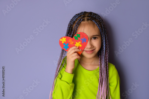 A girl with blue-purple Afro pigtails in a light green top holds a heart with puzzle pieces in her hands as a sign of solidarity with people suffering from autism spectrum disorder. World Autism day photo