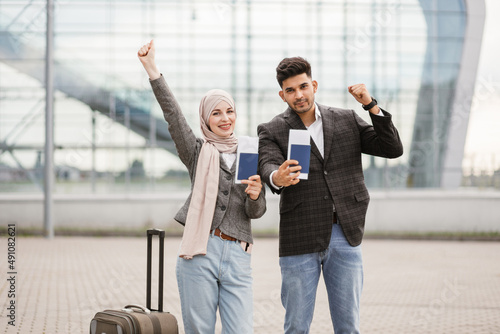 Smiling Muslim woman in hijab and Arab man, posing on camera outside modern airport terminal, demonstrating their passports and tickets while waiting flight. Ready for trip. Business people