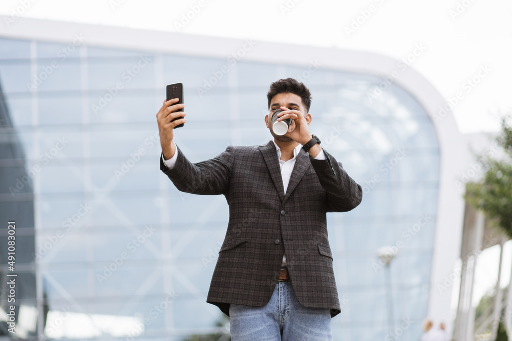 Cheerful good-looking Arabian businessman standing outdoors at new modern building, looking at mobile phone, making different emotions and gestures for selfie photo Cheerful good-looking Arabian businessman standing outdoors at new modern building, looking at mobile phone, making different emotions and gestures for selfie photo