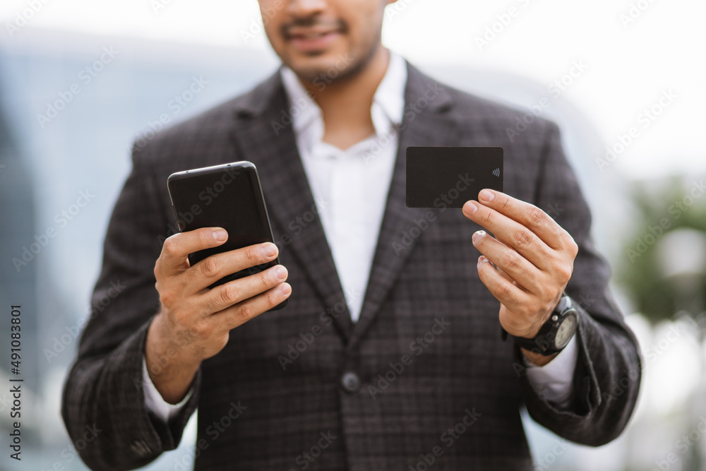 Business trip concept. Handsome young Indian man in eyeglasses, using smart phone and holding credit card with shopping online. Male freelancer receives payment online Business trip concept. Handsome young Indian man in eyeglasses, using smart phone and holding credit card with shopping online. Male freelancer receives payment online