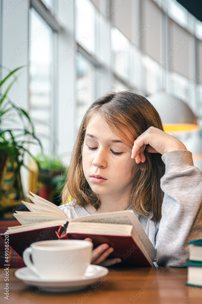 A little girl is reading a book while sitting in a cafe with a cup of tea. A little girl is reading a book while sitting in a cafe with a cup of tea.