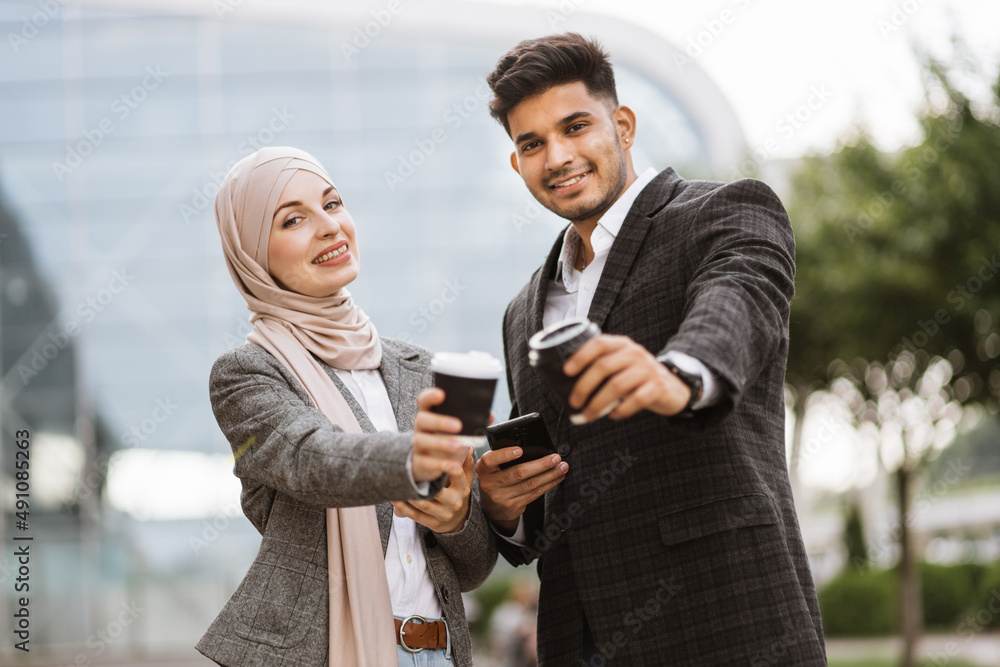 Smiling business couple, handsome Arab man and Muslim woman in hijab, standing outdoors in the street on coffee break, using phones and drinking take away coffee Smiling business couple, handsome Arab man and Muslim woman in hijab, standing outdoors in the street on coffee break, using phones and drinking take away coffee