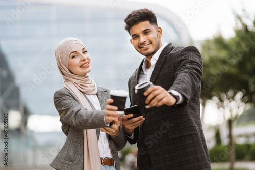 Smiling business couple, handsome Arab man and Muslim woman in hijab, standing outdoors in the street on coffee break, using phones and drinking take away coffee