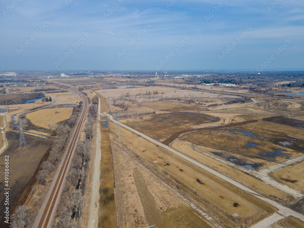 Aerial view of rural farm land with railroad tracks running through, dirt roads, blue sky, puddling water from melting snow. Trees along the railroad tracks. Transmission towers in the farm fields.  