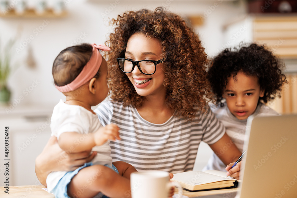 Obraz premium Young happy african american woman freelancer working remotely from home with two little kids