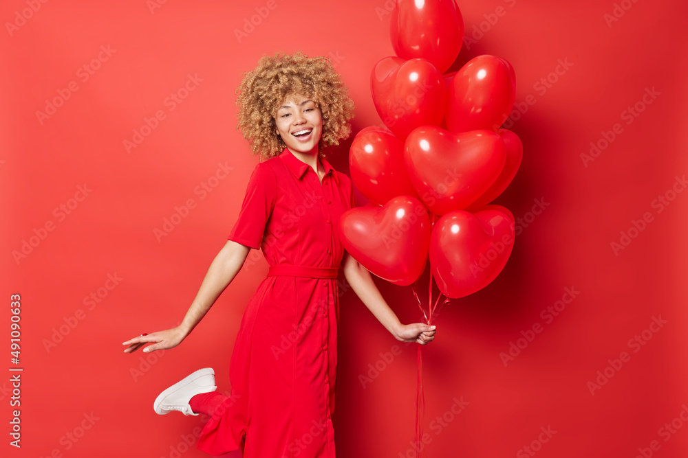 Joyful pretty woman dances with heart balloons enjoys celebration and party wears long dress smiles broadly expresses positive emotions isolated over red background. Happy Valentines Day concept Joyful pretty woman dances with heart balloons enjoys celebration and party wears long dress smiles broadly expresses positive emotions isolated over red background. Happy Valentines Day concept