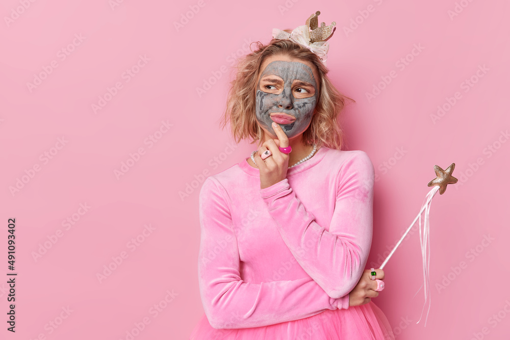 Dissatisfied young woman with beauty mask on face looks seriusly away thinks deeply about something considers how to organise party or holiday for childern wears festive dress poses against pink wall Dissatisfied young woman with beauty mask on face looks seriusly away thinks deeply about something considers how to organise party or holiday for childern wears festive dress poses against pink wall