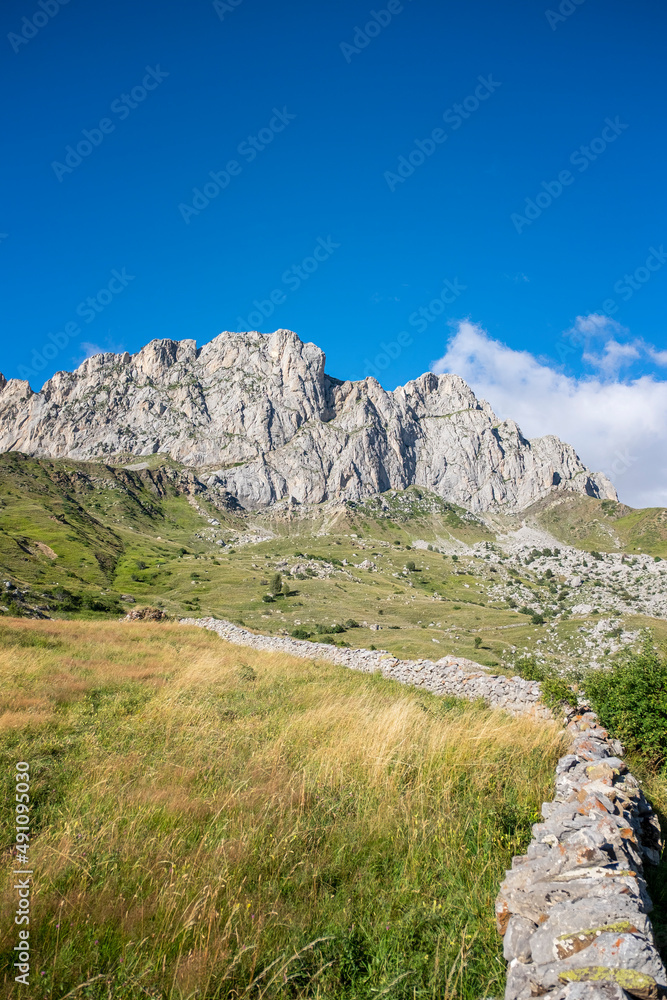 green meadows with grass at the foot of a rocky mountain on a sunny summer day, a stone wall out of focus in the foreground, Formigal, Huesca, Spain