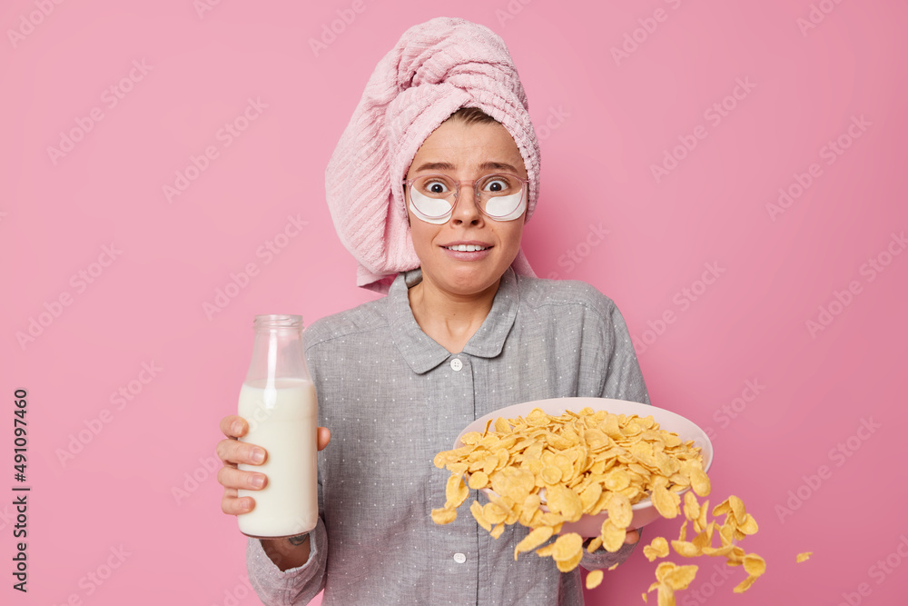 Stressed young woman looks embarrassed poses with cornflakes spilled from bowl holds bottle of milk wears pajama and bath towel wrapped over head poses against pink background. Time for breakfast Stressed young woman looks embarrassed poses with cornflakes spilled from bowl holds bottle of milk wears pajama and bath towel wrapped over head poses against pink background. Time for breakfast