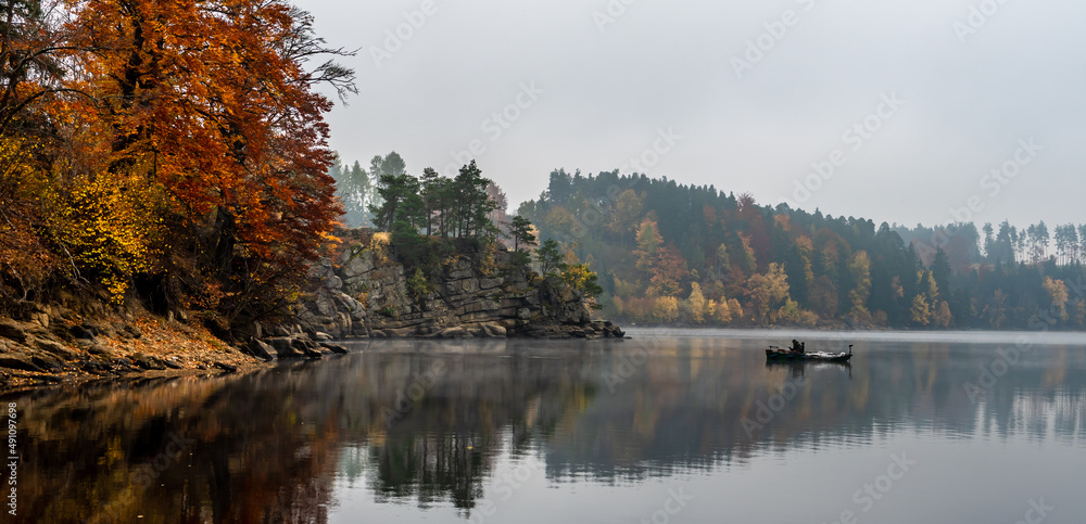 Fototapeta premium Foggy Landscape With Fishermans Boat On Calm Lake And Autumnal Forest At Lake Ottenstein In Austria
