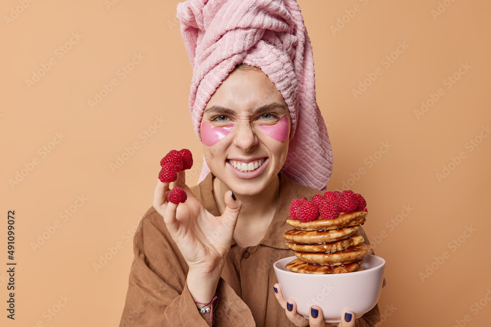 Positive young woman smirks face and shows white teeth has fun while eating delicious breakfast has raspberries on fingers wears wrapped towel on head pajama applies beauty patches under eyes Positive young woman smirks face and shows white teeth has fun while eating delicious breakfast has raspberries on fingers wears wrapped towel on head pajama applies beauty patches under eyes