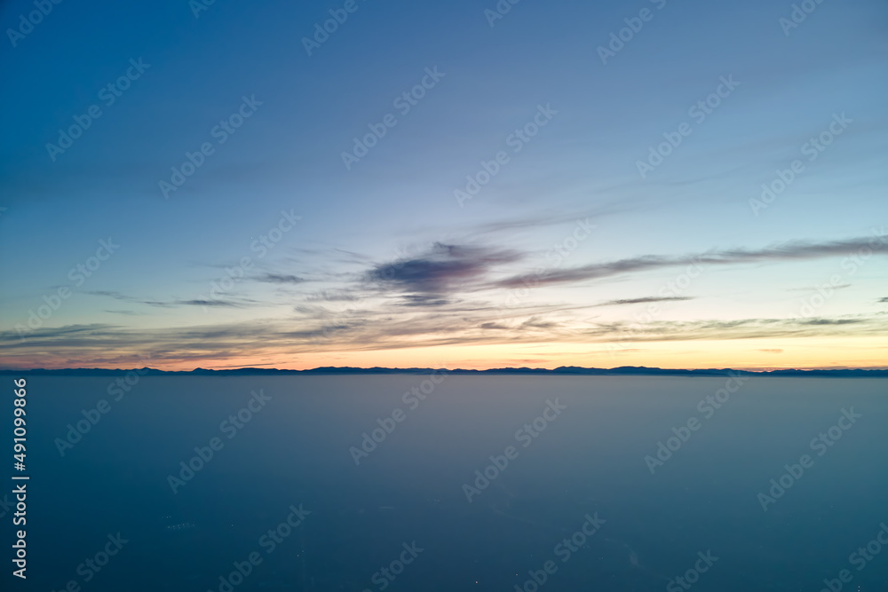 Aerial view of colorful sunset over white dense foggy clouds cover with distant dark silhouettes of mountain hills on horizon