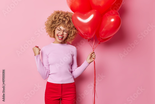 Horizontal shot of overjoyed curly haired woman clenches fist celebrates success or good news holds bunch of red heart balloons has upbeat mood dressed casually isolated over pink background.