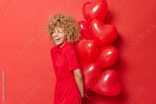 Overjoyed curly haired young woman laughs out happily holds big bunch of heart balloons behind back prepares surprise for lover on Valentines Day wears dress isolated over vivid red background.