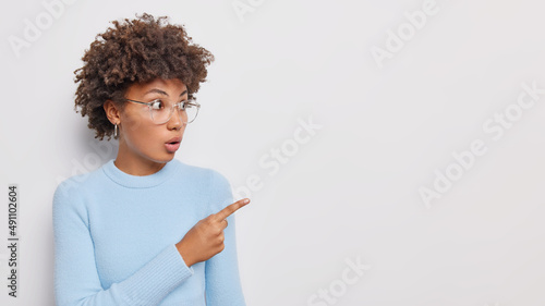 Impressed stupefied young woman points index finger away shows place for your advertisement reacts on big discounts wears eyeglasses and casual jumper isolated over white background. Omg look there