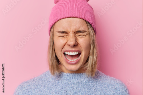 Headshot of emotional irritated woman screams loudly keeps mouth opened feels angry wears hat and sweater expresses negative emotions isolated over pink background. Outraged girl yelling from anger