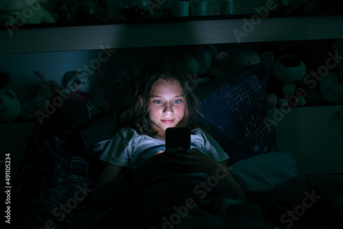 Obraz na płótnie A little girl lies in bed at night and types on a glowing smartphone screen. w salonie