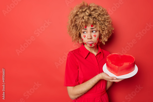 Upset curly haired young woman has sad expression holds heart cake looks away wears dress feels lonely on Valentines Day isolated over red background blank space for your promotional content