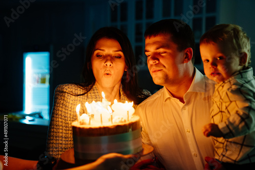 a father and mother with a kid son make a wish and blow out candles on the cake.