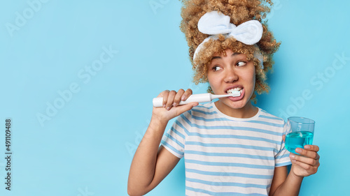 Horizontal shot of curly haired young woman brushes teeth with electric toothbrush uses mouthwash for fresh breath wears headband and casual striped t shirt isolated over blue background copy space photo