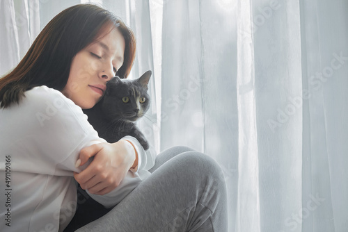Photography A beautiful young girl sits alone at the window at home and is bored, she is sad, she holds a dark gray cat with bright eyes in her arms