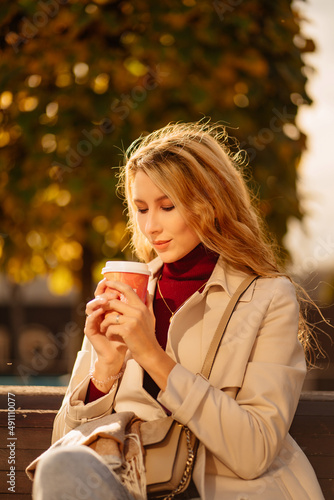 a beautiful blonde with a cup of coffee sits on a bench in an autumn park.