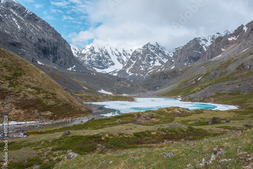 Atmospheric mountain landscape with frozen alpine lake and high snowy mountains under cloudy sky. Awesome highland scenery with icy mountain lake on background of large snow mountains in low clouds.