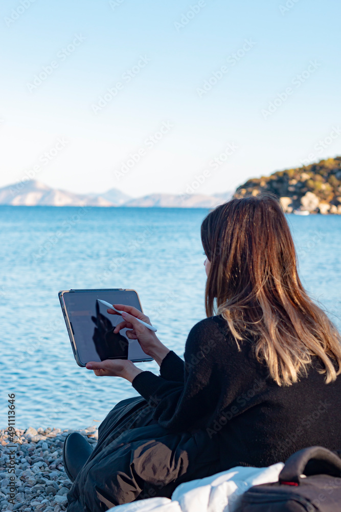 Back view of young girl drawing on tablet with pen by sea. Her face is not see. Unrecognizable person. Creativity and design concept. Vertical photo.