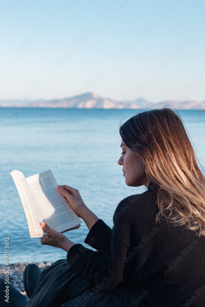 Back view of young girl reading a book by sea. Her face is not seen. Unrecognizable person. 