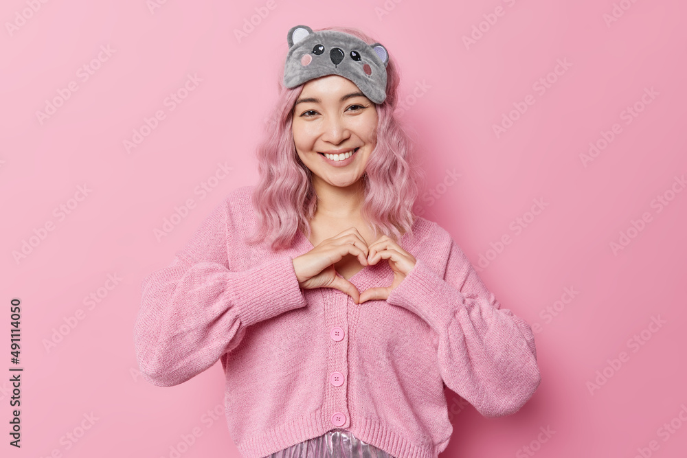 Positive young pink haired Asian girl makes heart gesture expresses love smiles pleasantly wears sleepmask on forehead and casual jumper isolated over pink background. Body language concept. Positive young pink haired Asian girl makes heart gesture expresses love smiles pleasantly wears sleepmask on forehead and casual jumper isolated over pink background. Body language concept.