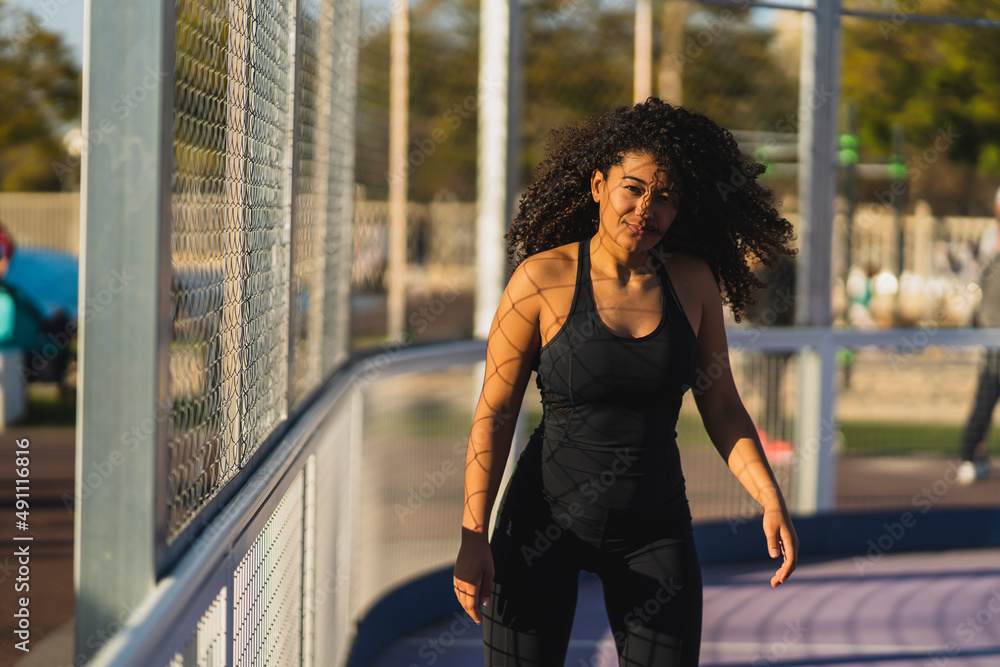 Chica marroquí morena con pelo largo rizado en patines posando junto a una verja de espacio deportivo habilitado Chica marroquí morena con pelo largo rizado en patines posando junto a una verja de espacio deportivo habilitado
